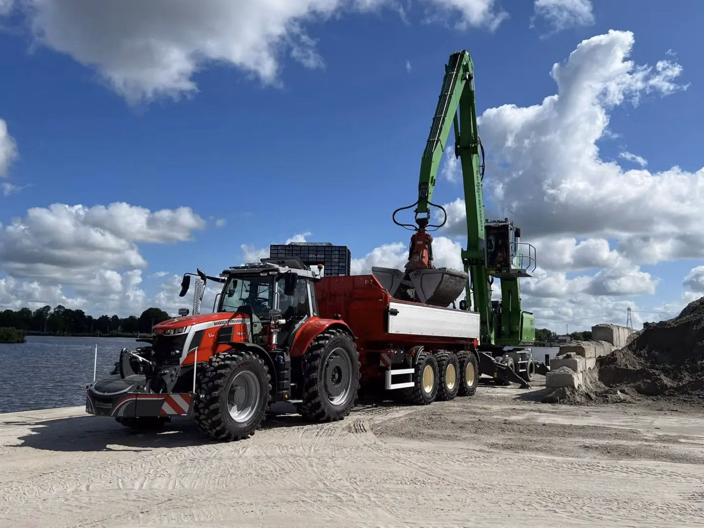 A red tractor with a front weight bumper is attached to a trailer being loaded by a green excavator by a river.