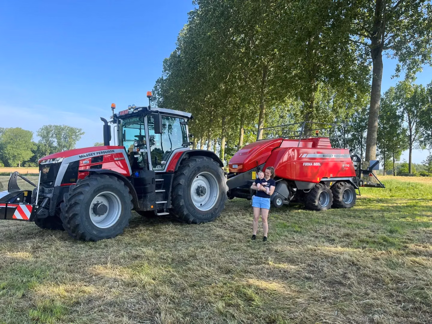 A red Massey Ferguson tractor with a front weight bumper attached, next to a baler in a grassy field with two people nearby