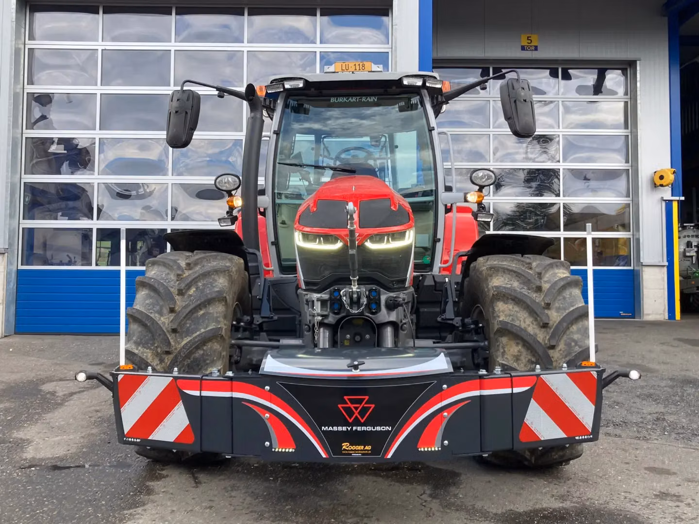 Red tractor with a Massey Ferguson front weight bumper featuring LED lights and reflective panels, parked outside a building