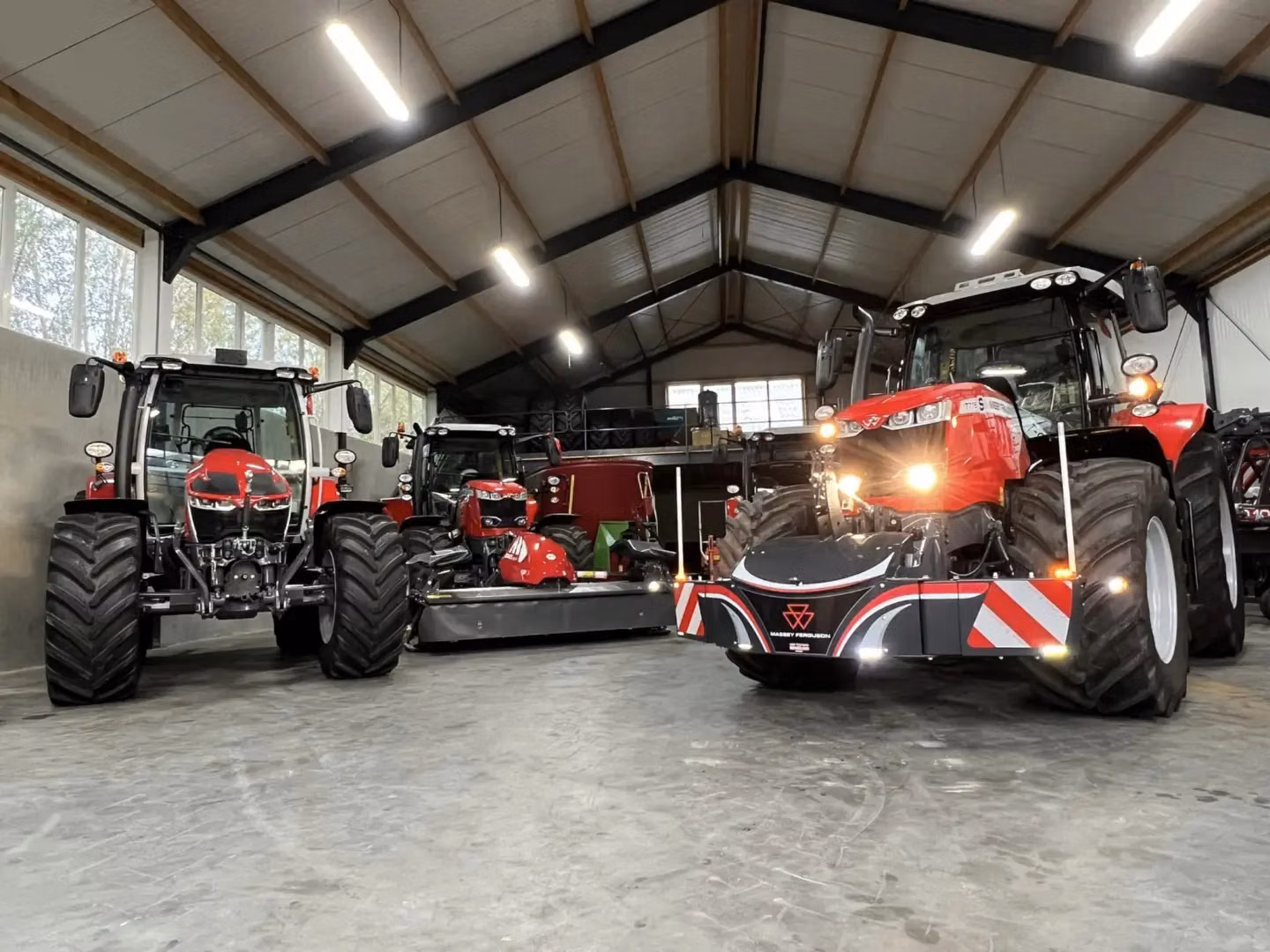 Three red tractors parked indoors, with the rightmost tractor fitted with a front weight bumper with LED lights and safety