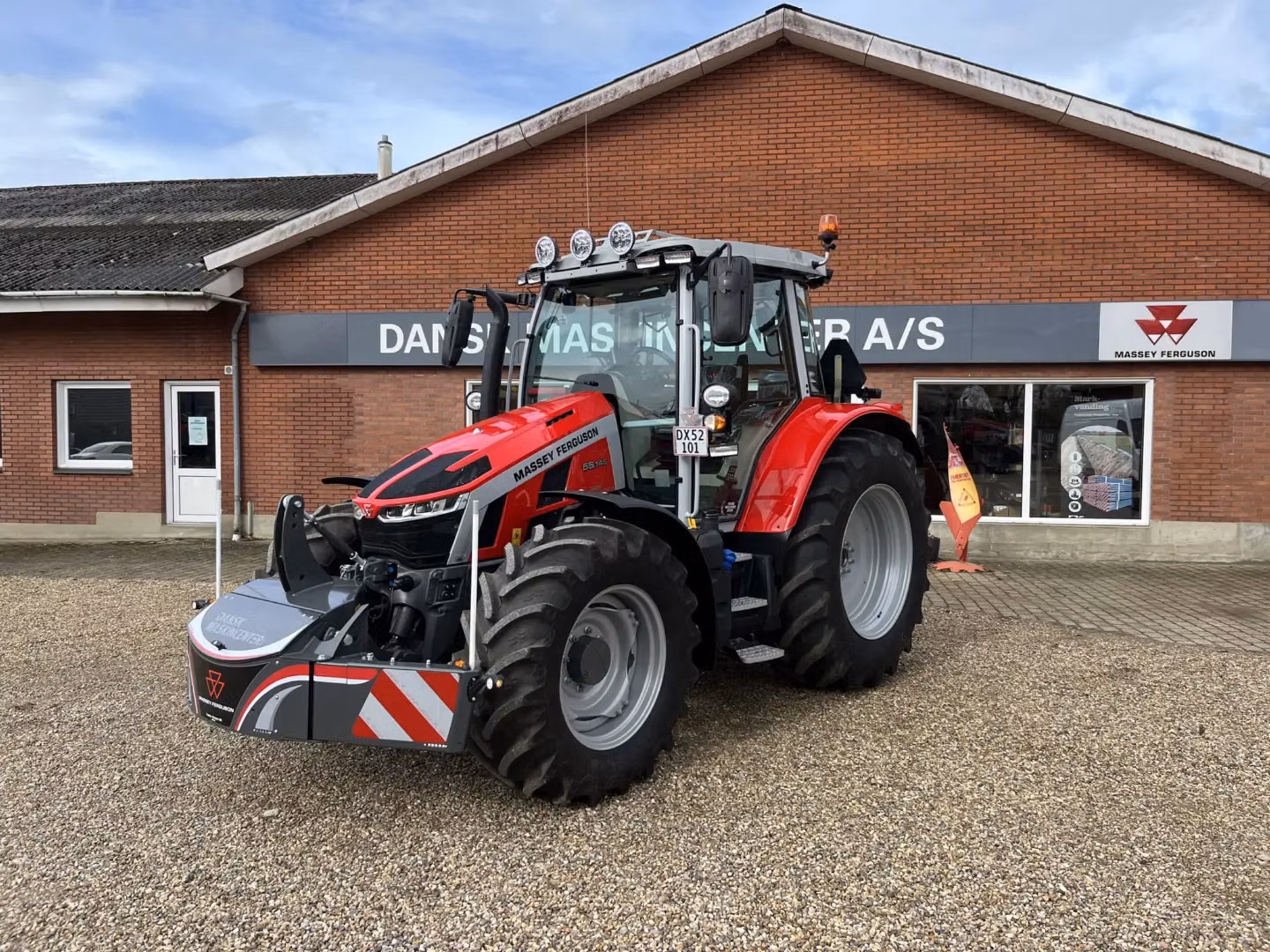 Red tractor with a front weight bumper featuring LED lights and safety markings, parked outside a brick building