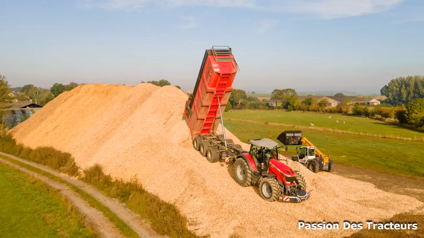 Red tractor with a front weight bumper and tipped trailer unloading wood chips beside a large pile in a rural field