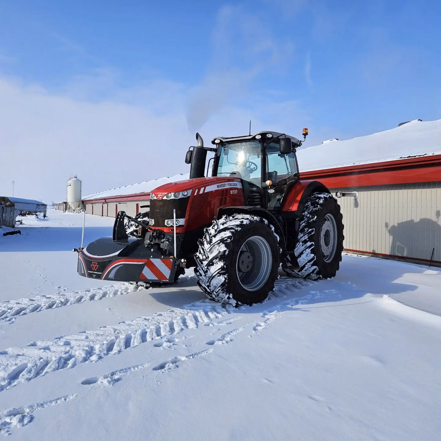 A red tractor with a front weight bumper and LED lights is parked on snowy ground beside a building.