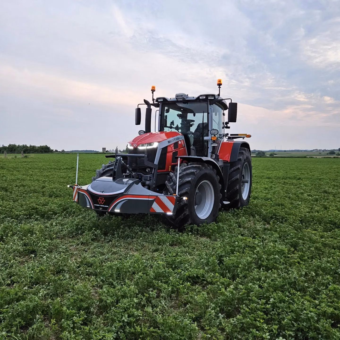 Red tractor with a front weight bumper featuring LED lights and red-white safety markings on a green field