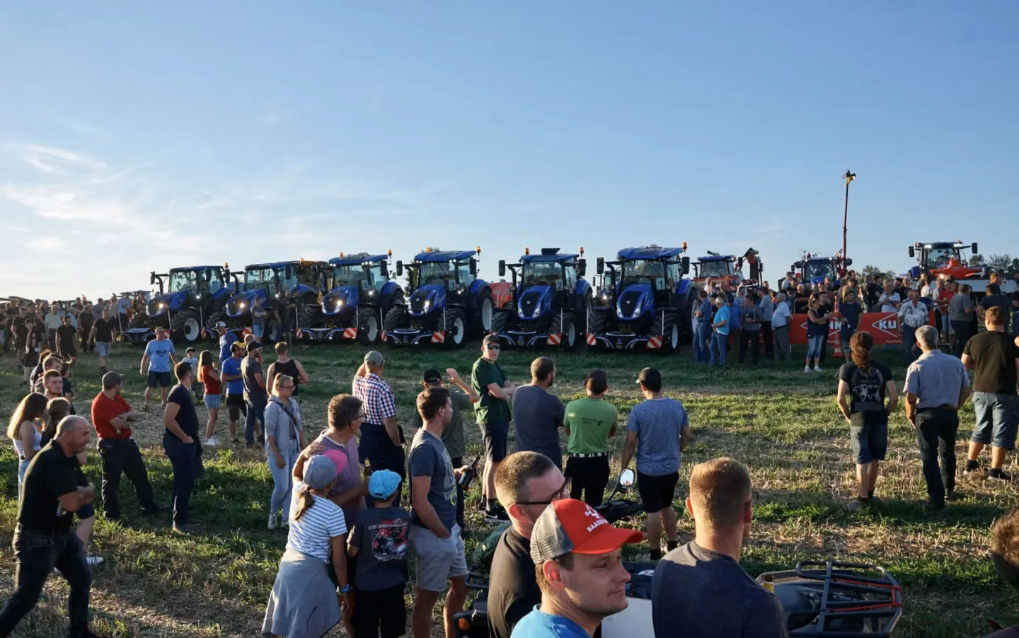 Several blue tractors fitted with front weight bumpers lined up in a field with a crowd of people gathered around