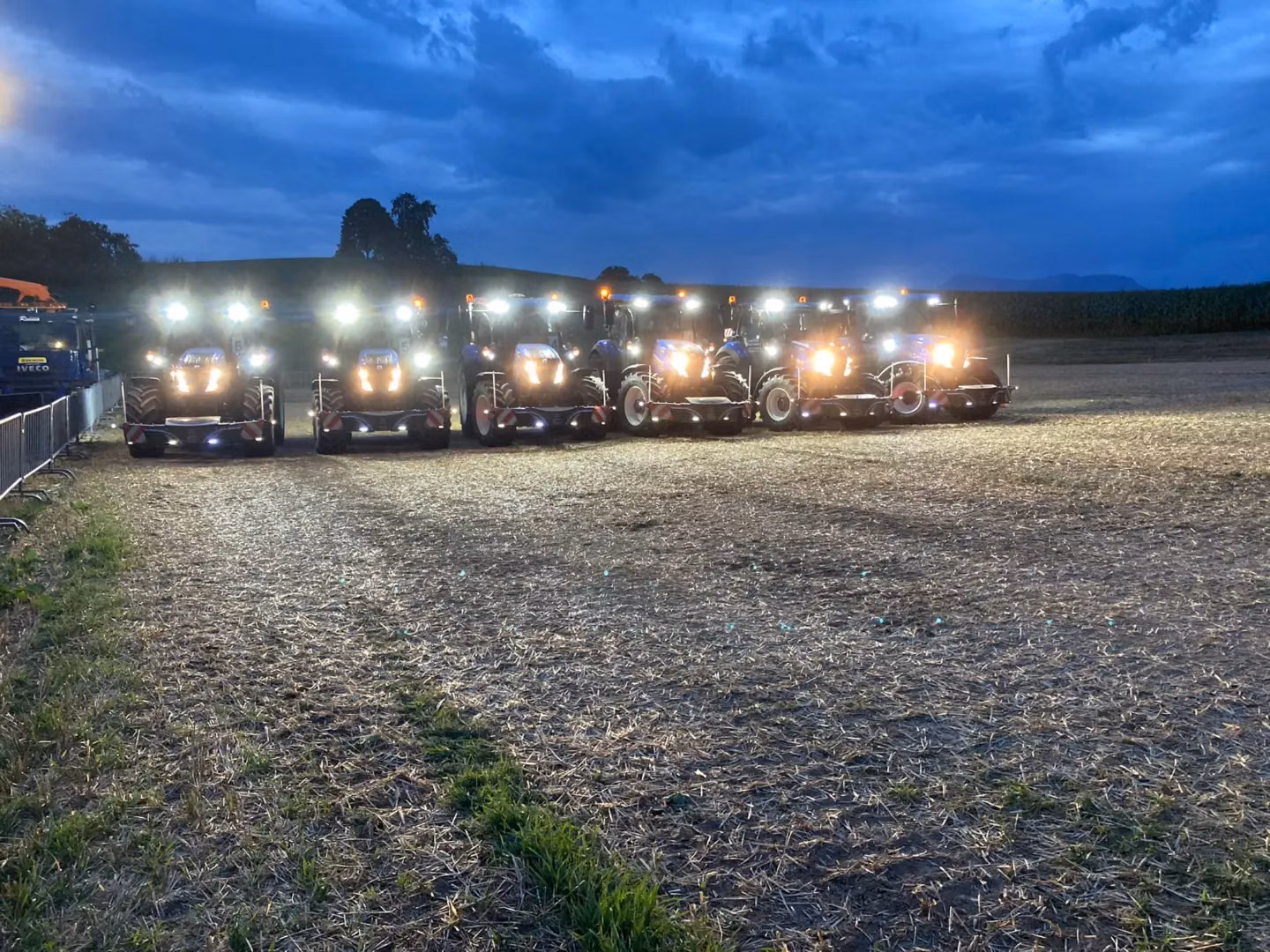 Seven tractors with front weight bumpers are lined up in a field at dusk with headlights shining brightly