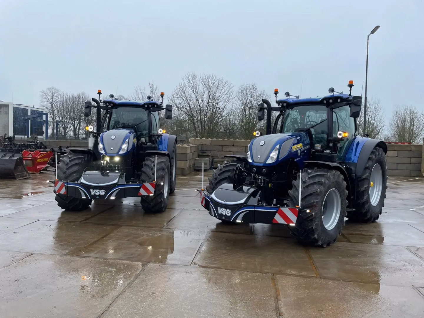 Two blue tractors are parked outside on wet concrete, each fitted with a large black front weight bumper marked VSG