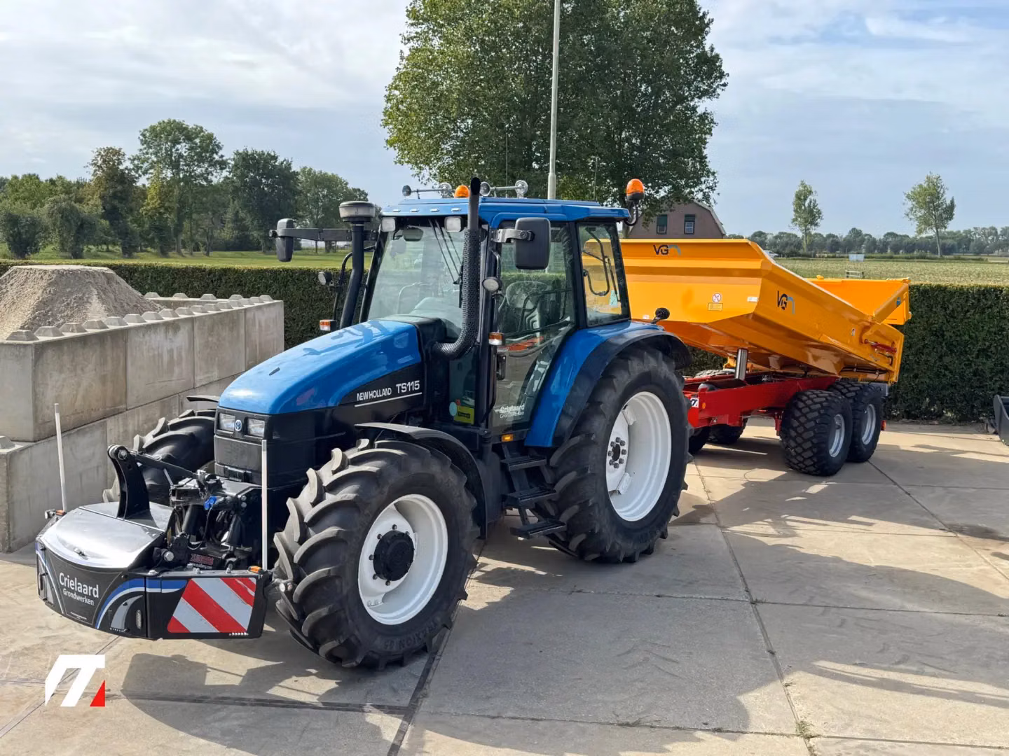 Blue tractor with a front weight bumper attached, hitched to a large yellow trailer, parked on a paved area