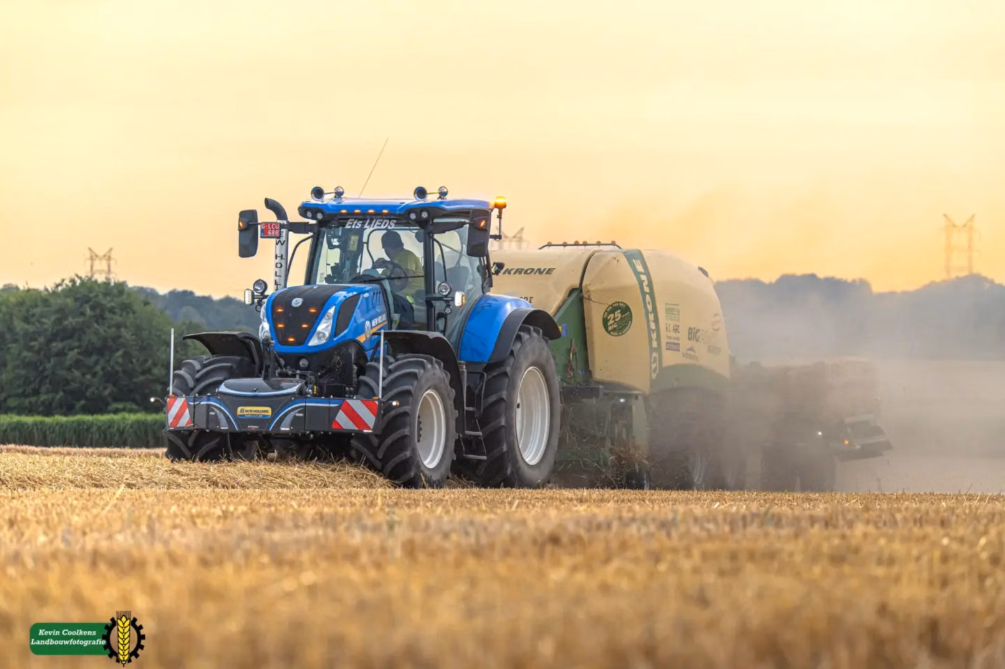 A blue tractor with a front weight bumper works in a straw field, pulling a large round baler behind it