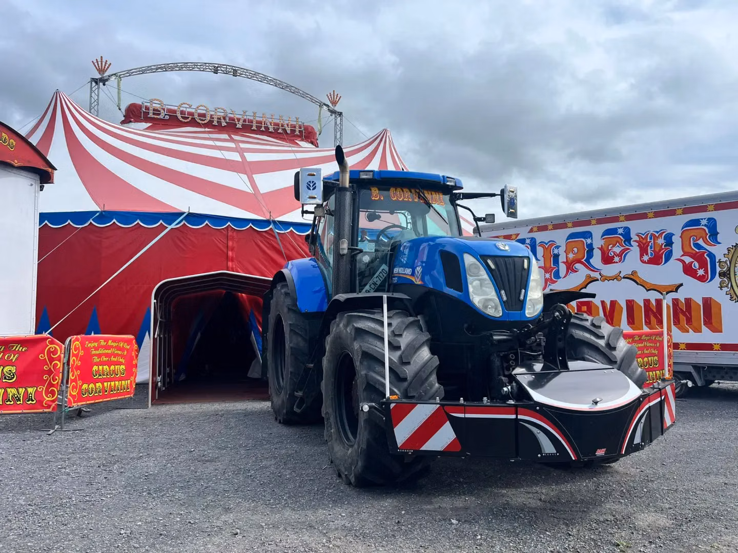A blue tractor with a black front weight bumper with reflective stripes parked in front of a circus tent and sign