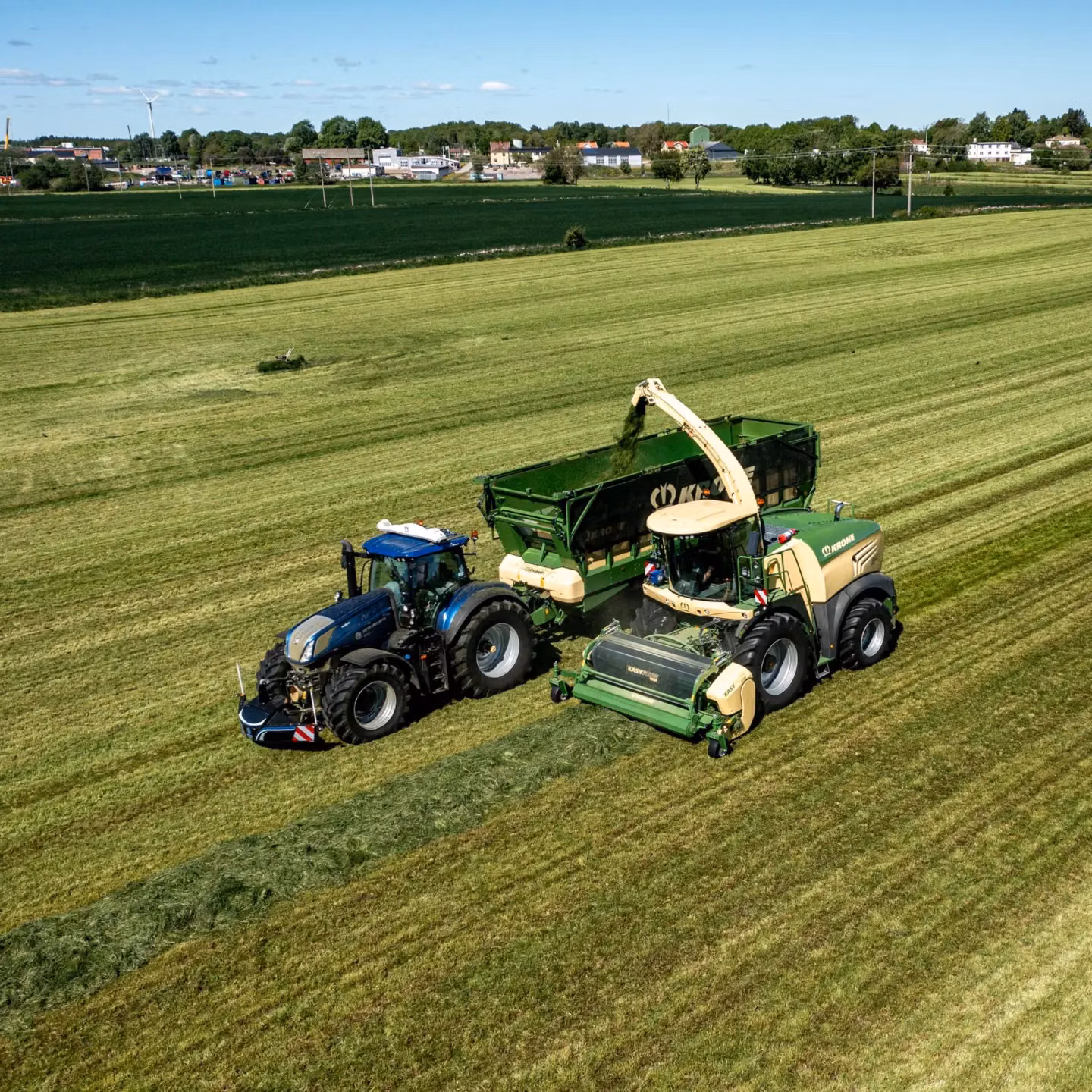 A blue tractor with a front weight bumper alongside a forage harvester loading green crop into a trailer in a field