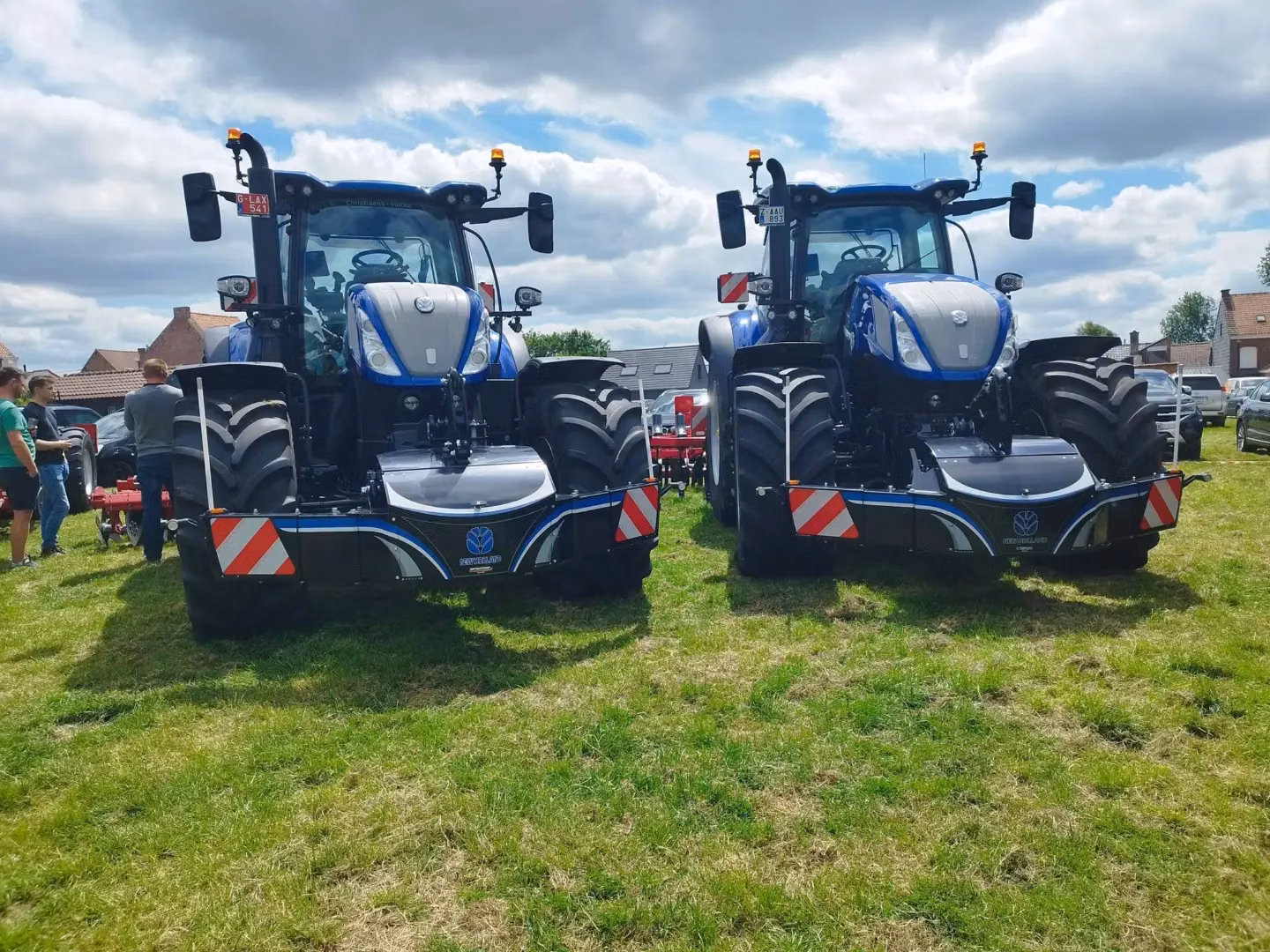 Two blue tractors with large front weight bumpers featuring LED lights are parked on grass with people nearby