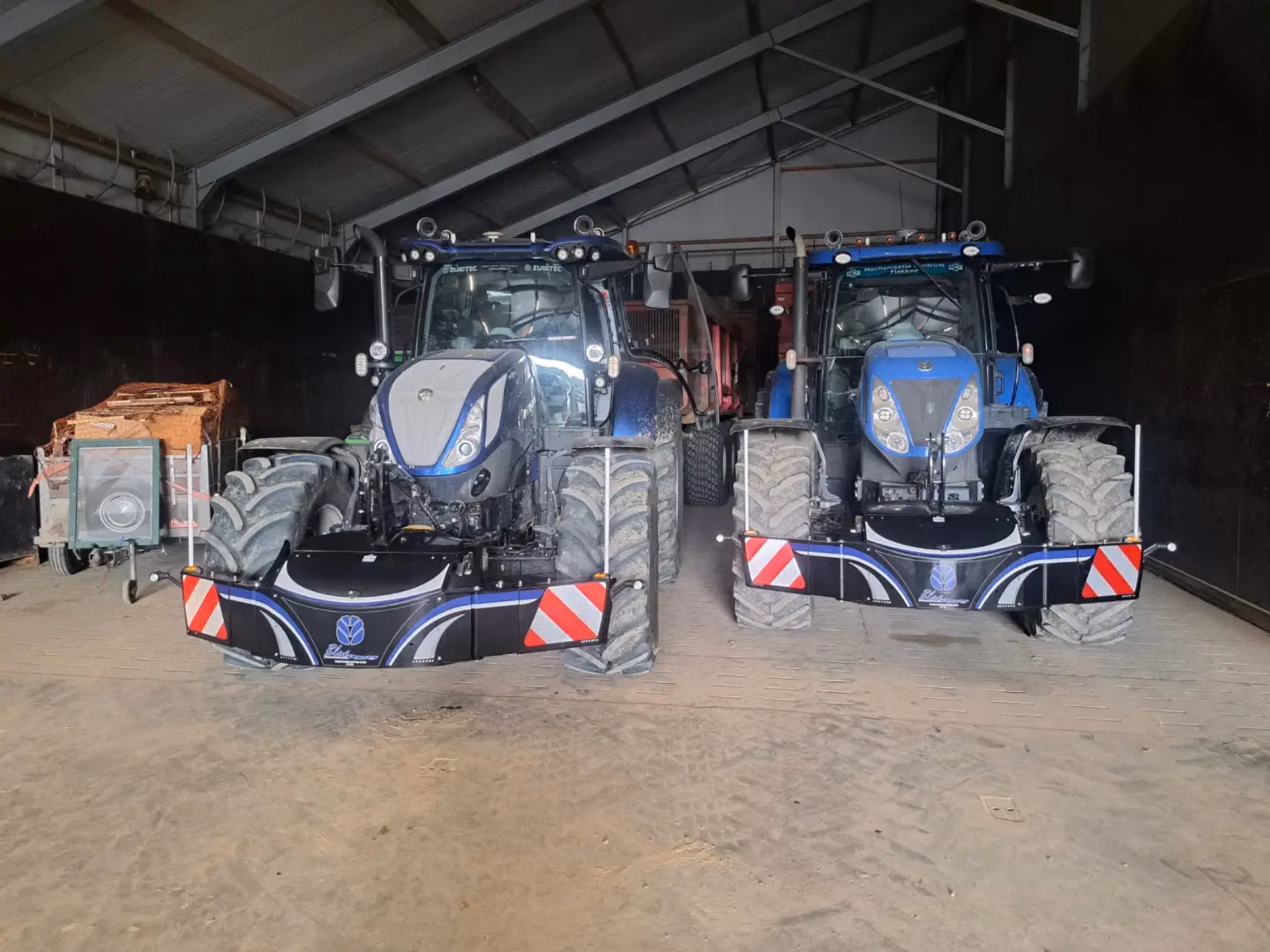 Two tractors parked in a shed, each fitted with a front weight bumper featuring reflective red and white stripes