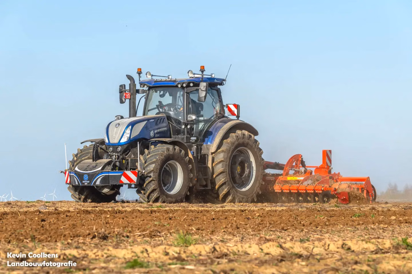 Blue tractor with a front weight bumper and rear-mounted orange soil cultivator working on a field