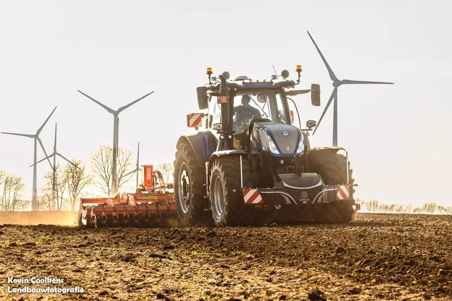 A blue tractor with a front weight bumper and LED lights works in a field with wind turbines in the background