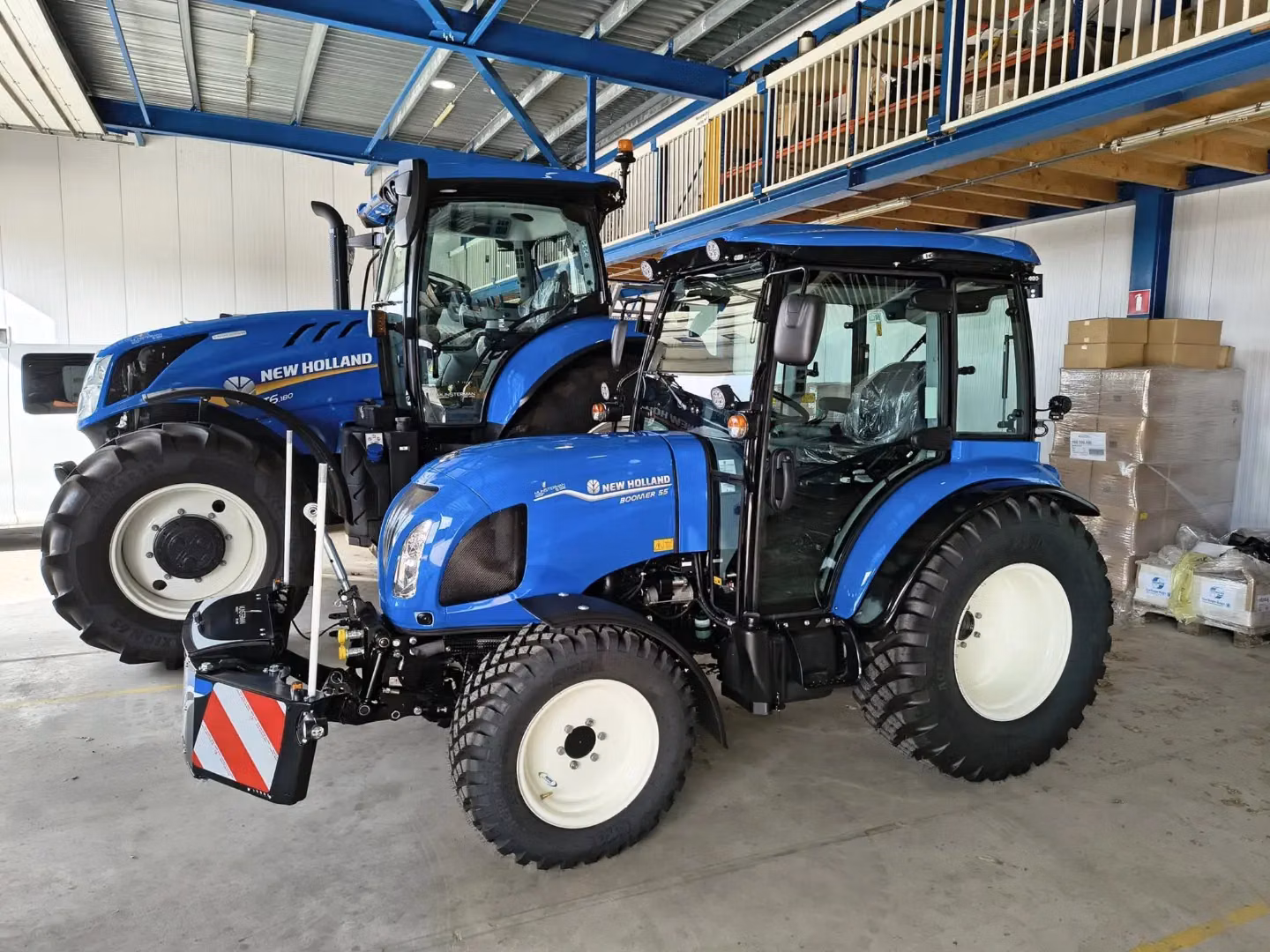 Two blue tractors indoors with the front one fitted with a black and red striped front weight bumper attachment