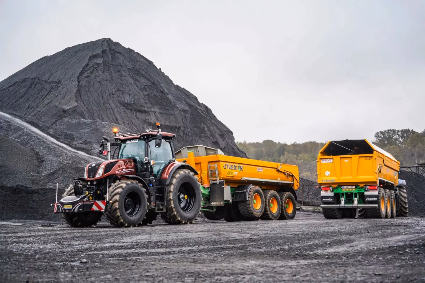 A tractor with a front weight bumper is hitched to a yellow trailer on a gravel surface near a large mound of dark material