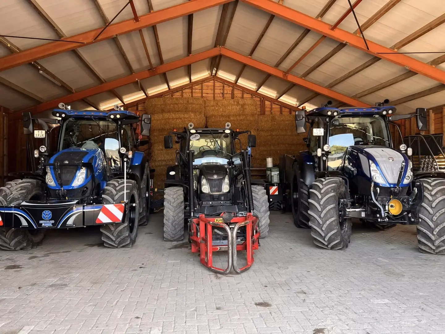 Three tractors parked in a shed with the left tractor fitted with a large front weight bumper marked with reflective stripes