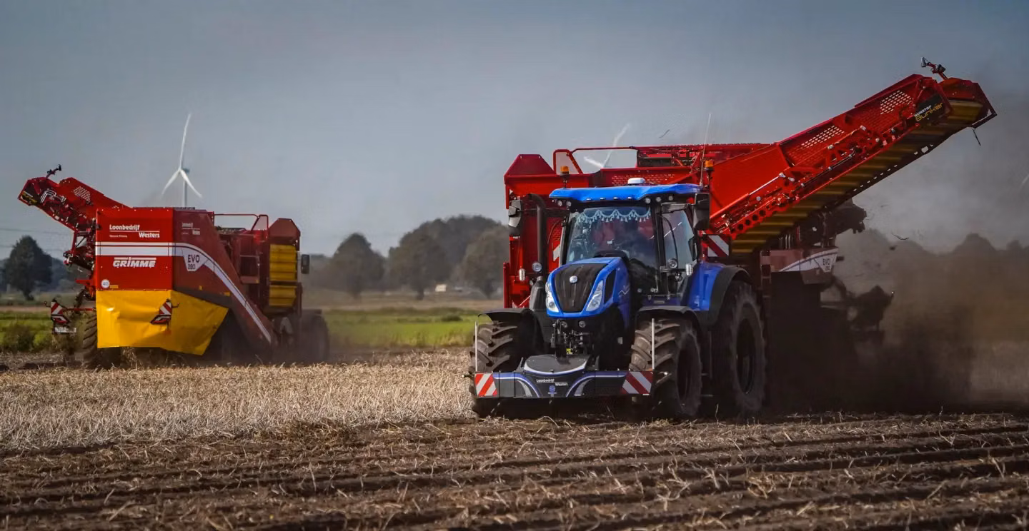 Blue tractor with a front weight bumper attached, working in a field with large red harvesting machinery