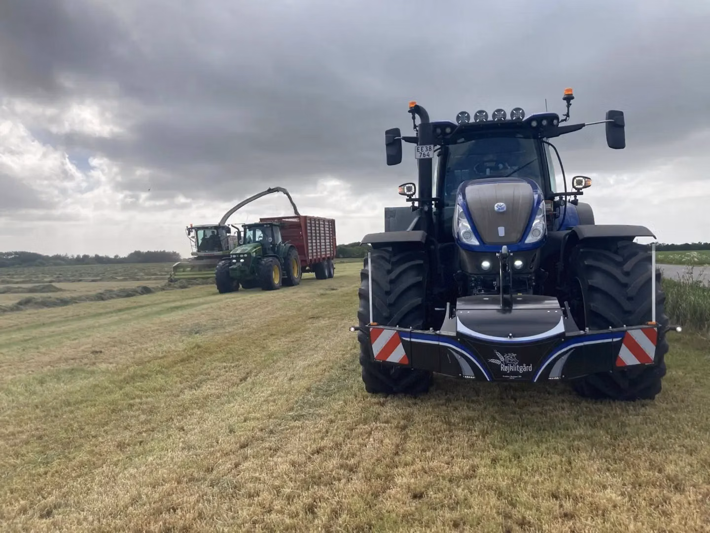 Blue tractor with a front weight bumper is parked on grass, with another tractor and forage harvester in the background