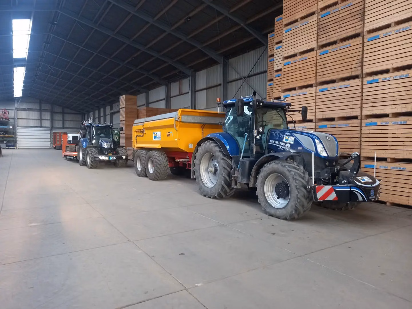 Blue tractor with a front weight bumper and yellow trailer parked inside a warehouse with stacked wooden crates