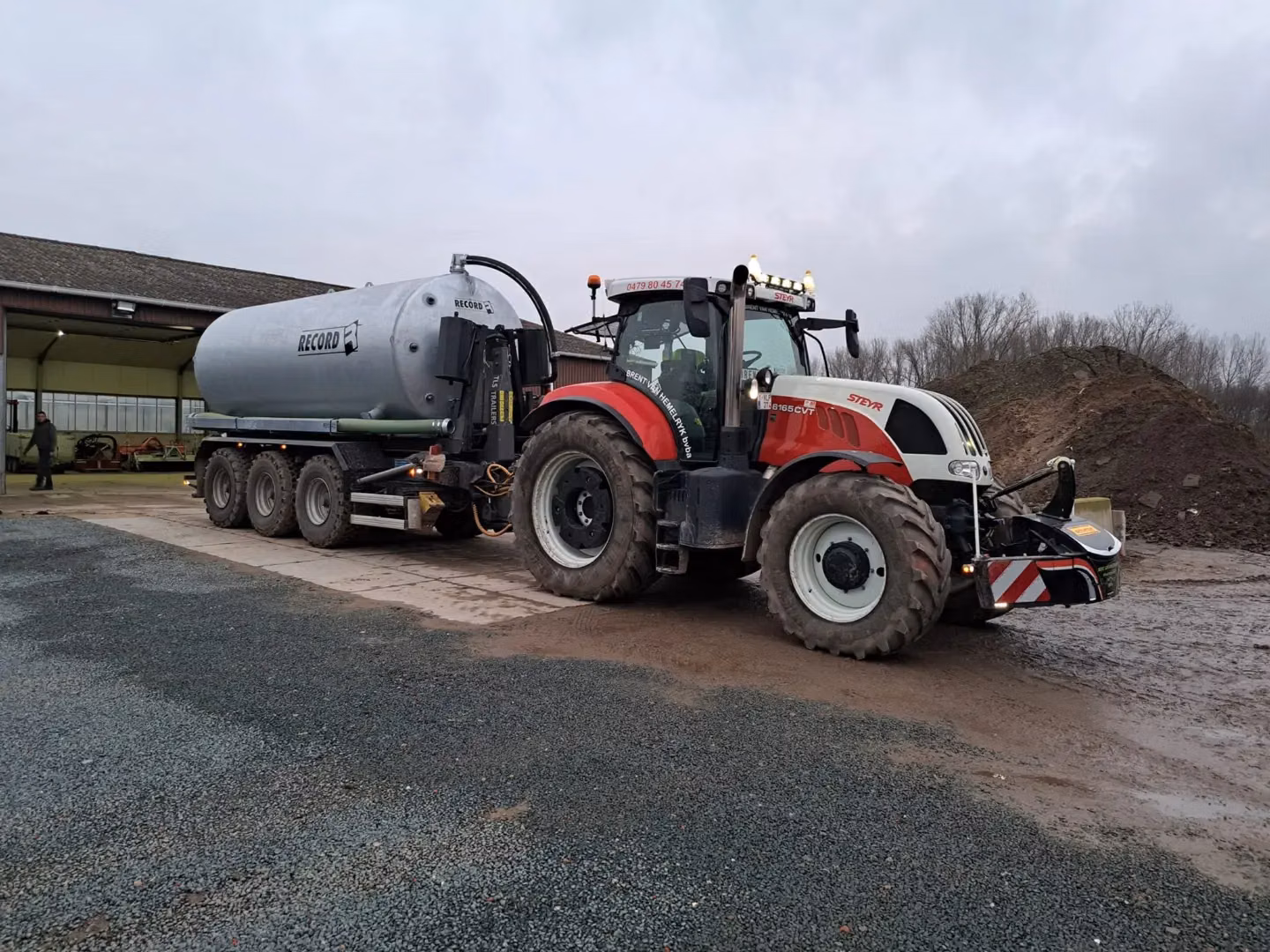 Red and white tractor with a front weight bumper attached, towing a large silver tank trailer on a muddy farmyard