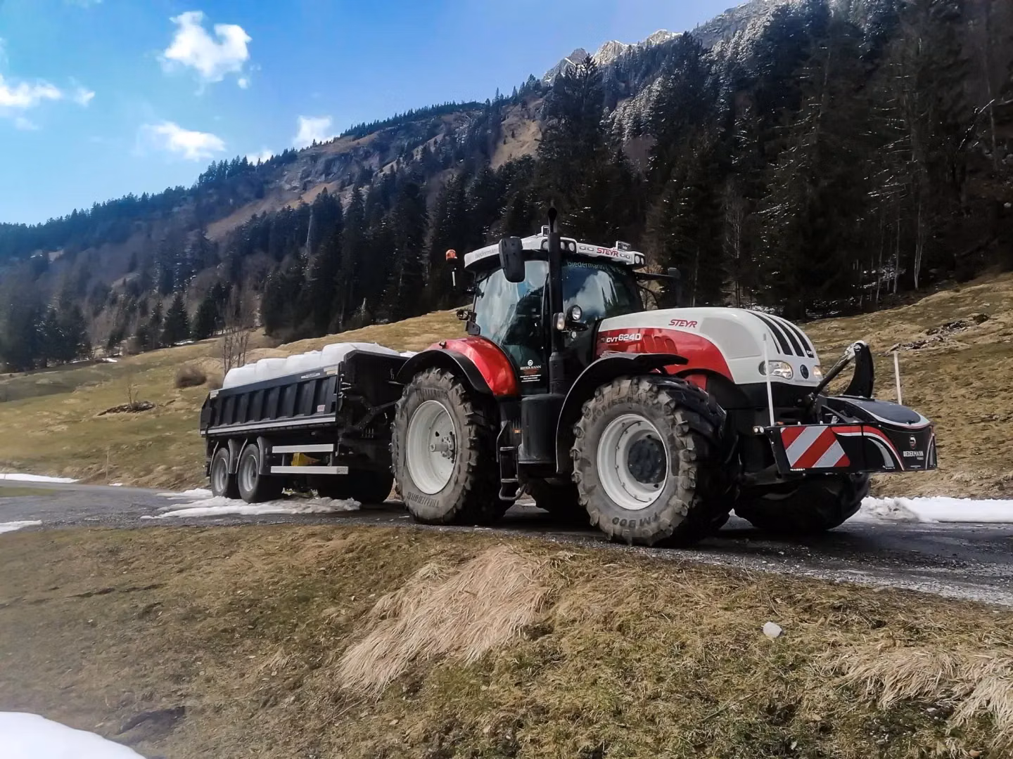 A red and white tractor with a front weight bumper is towing a trailer on a partially snow-covered rural road