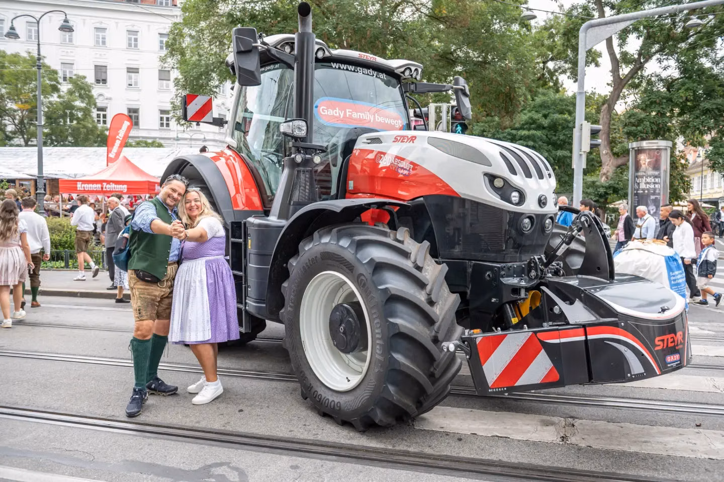 A man and woman in traditional clothing pose smiling in front of a Steyr tractor with a front weight bumper