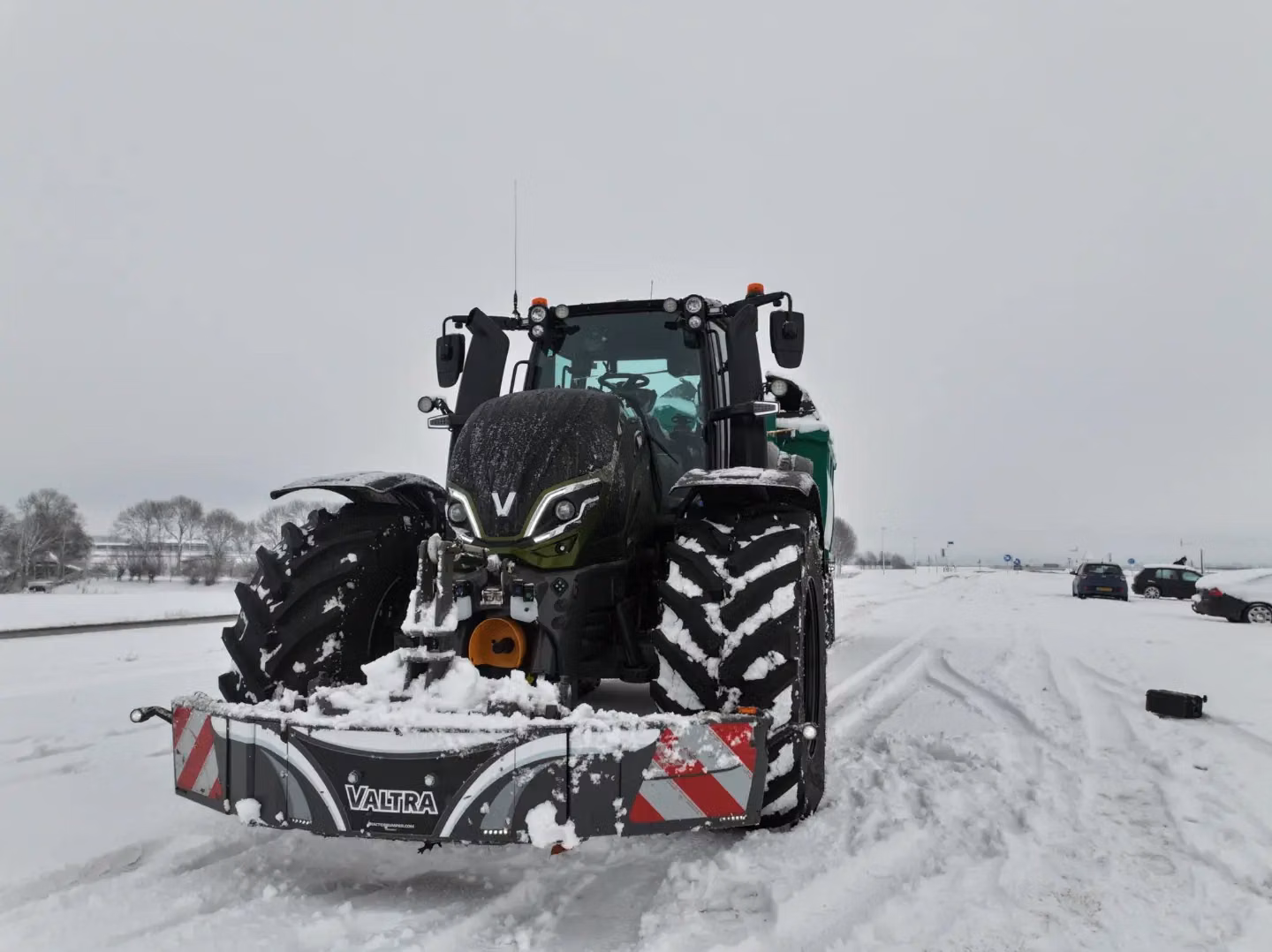 A tractor with a Valtra front weight bumper is parked on a snowy road with trees and cars in the background