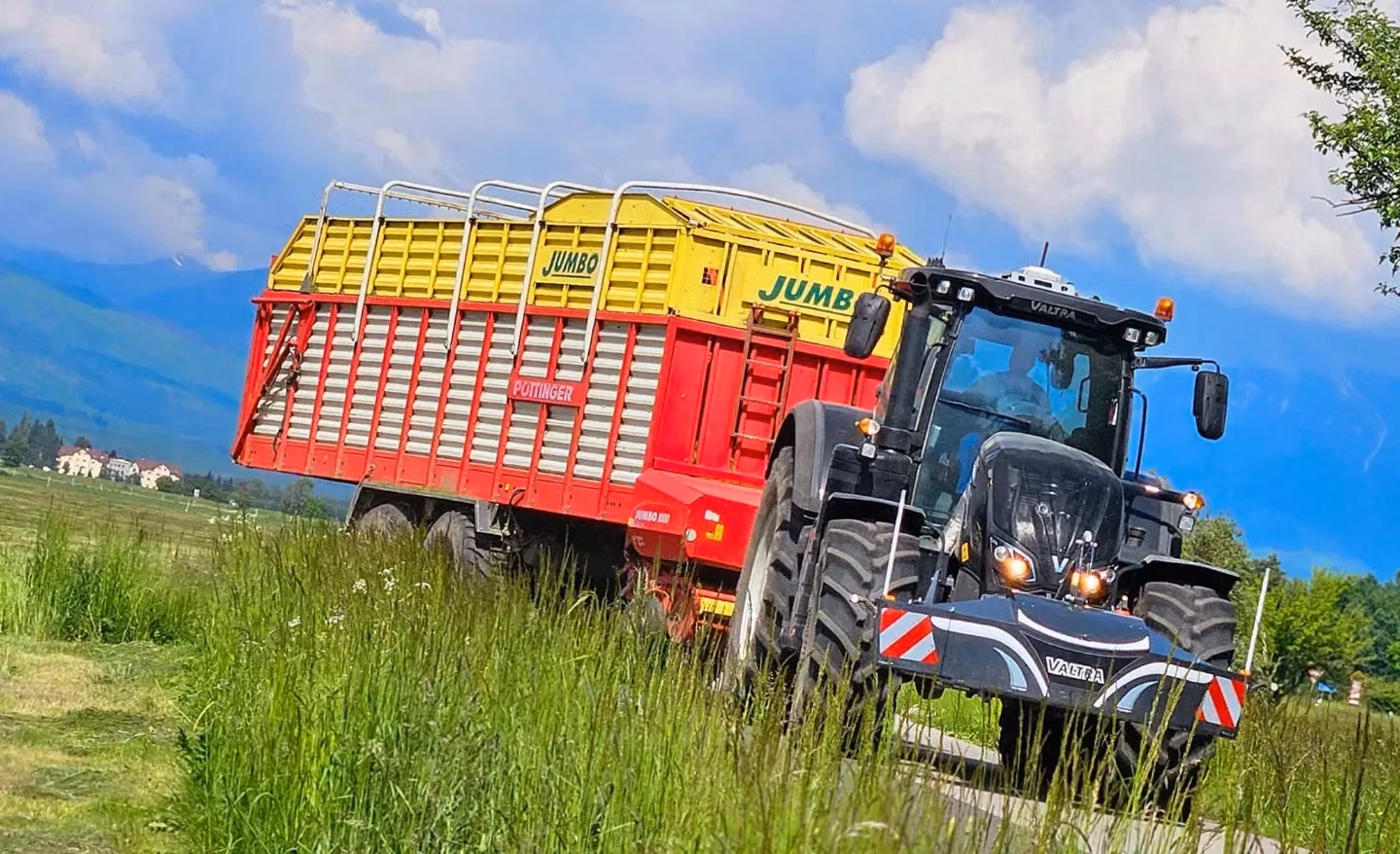 A black tractor with a front weight bumper and LED lights pulls a large red and yellow trailer on a rural road