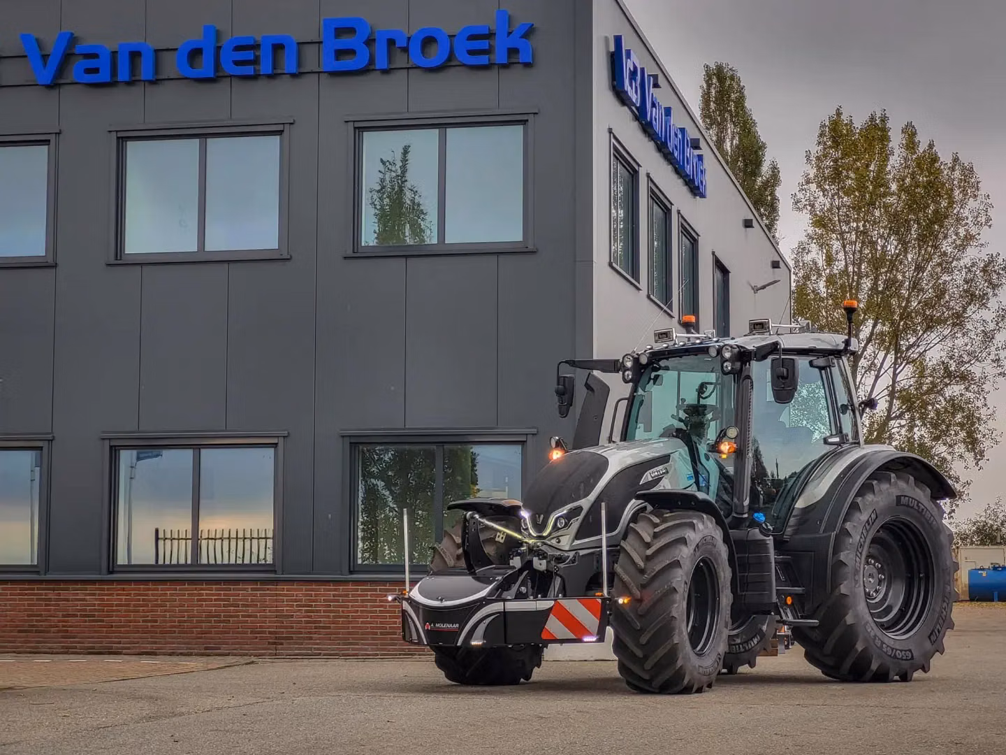 A black tractor with a front weight bumper and LED lights is parked in front of a building with large windows
