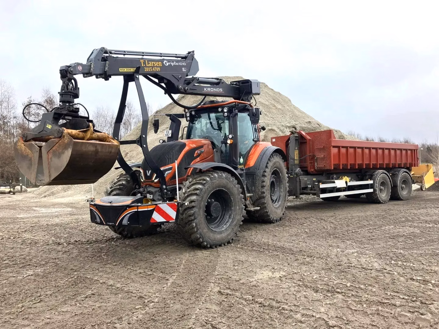 Tractor with a front weight bumper and grab attachment, towing a red trailer on a dirt worksite