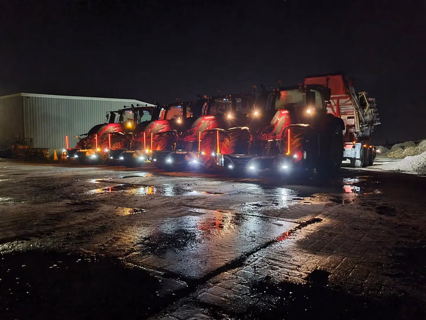 Five tractors at night with illuminated front weight bumpers lined up on a wet, reflective industrial surface