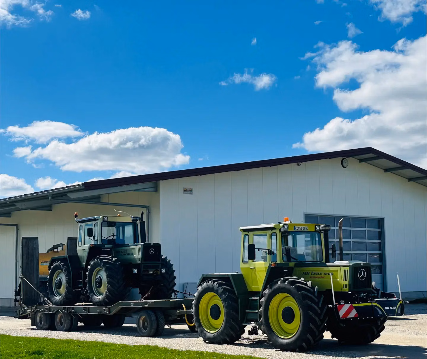 Two green tractors, one on a trailer and one parked in front, both with front weight bumpers, outside a building