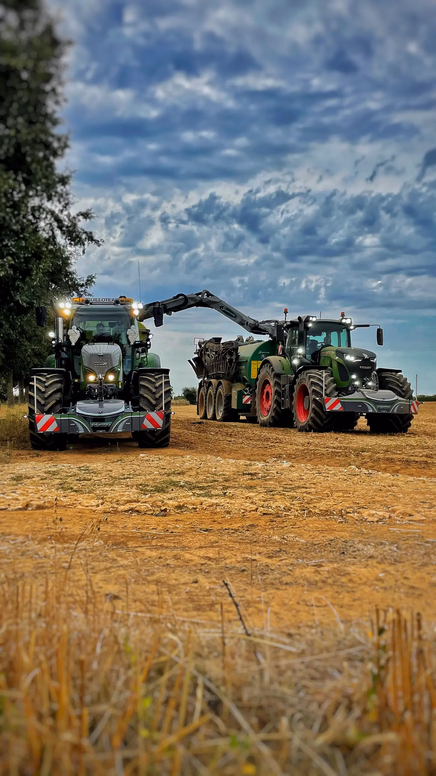 Two large green tractors fitted with front weight bumpers are working in a dry, open field under a cloudy sky