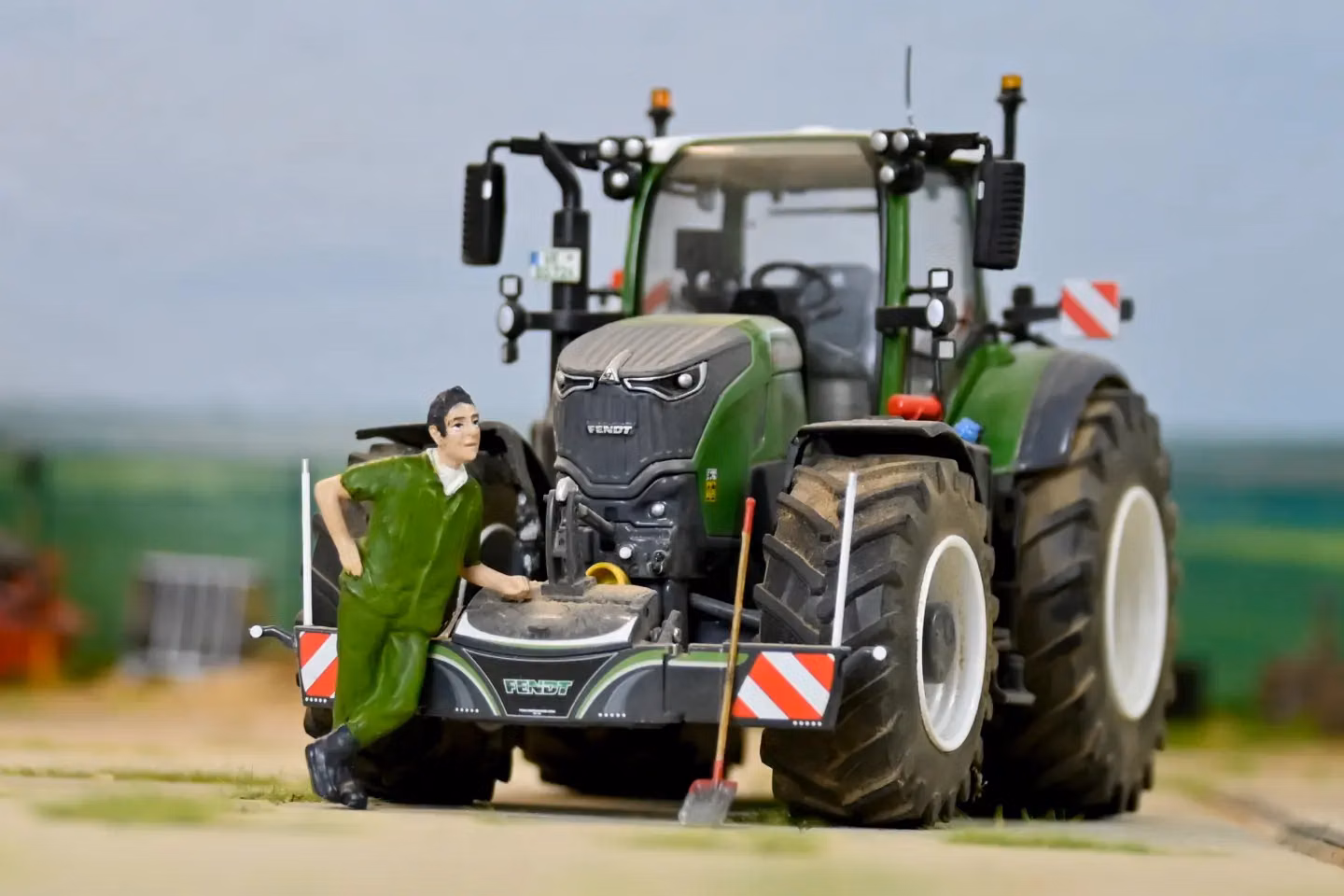 A model tractor with a front weight bumper, warning stripes and a figurine in green overalls leaning on it