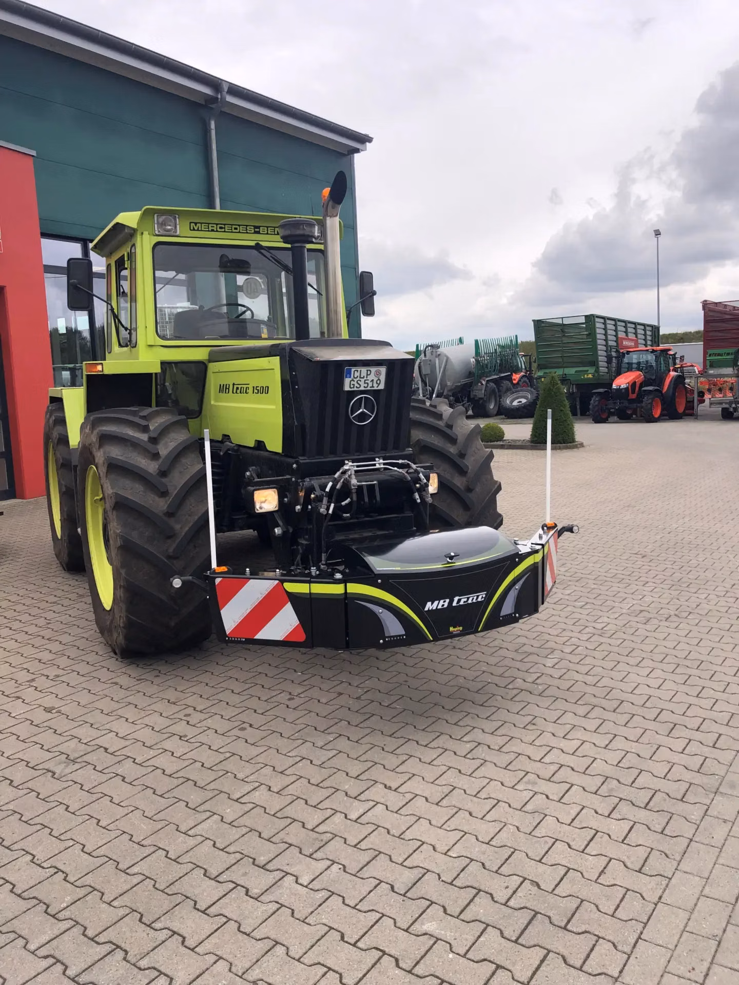 A large green tractor with a black front weight bumper featuring reflective stripes and MB trac branding attached.
