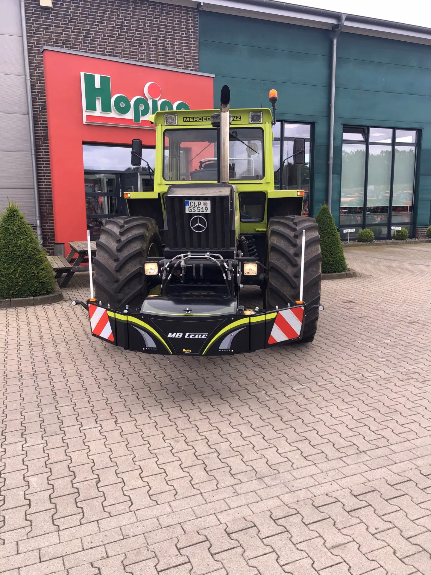 A tractor fitted with an MB trac front weight bumper featuring reflective stripes, parked on paving outside a building.
