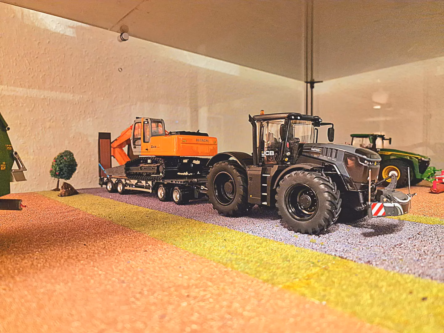 A miniature JCB tractor with a front weight bumper is parked near a flatbed trailer carrying an orange excavator.