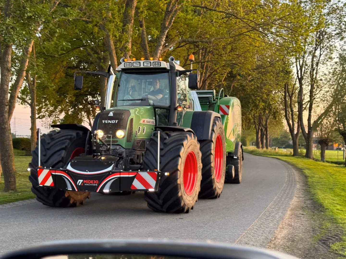 Green tractor with a visible front weight bumper driving on a rural road lined with trees, pulling a green trailer