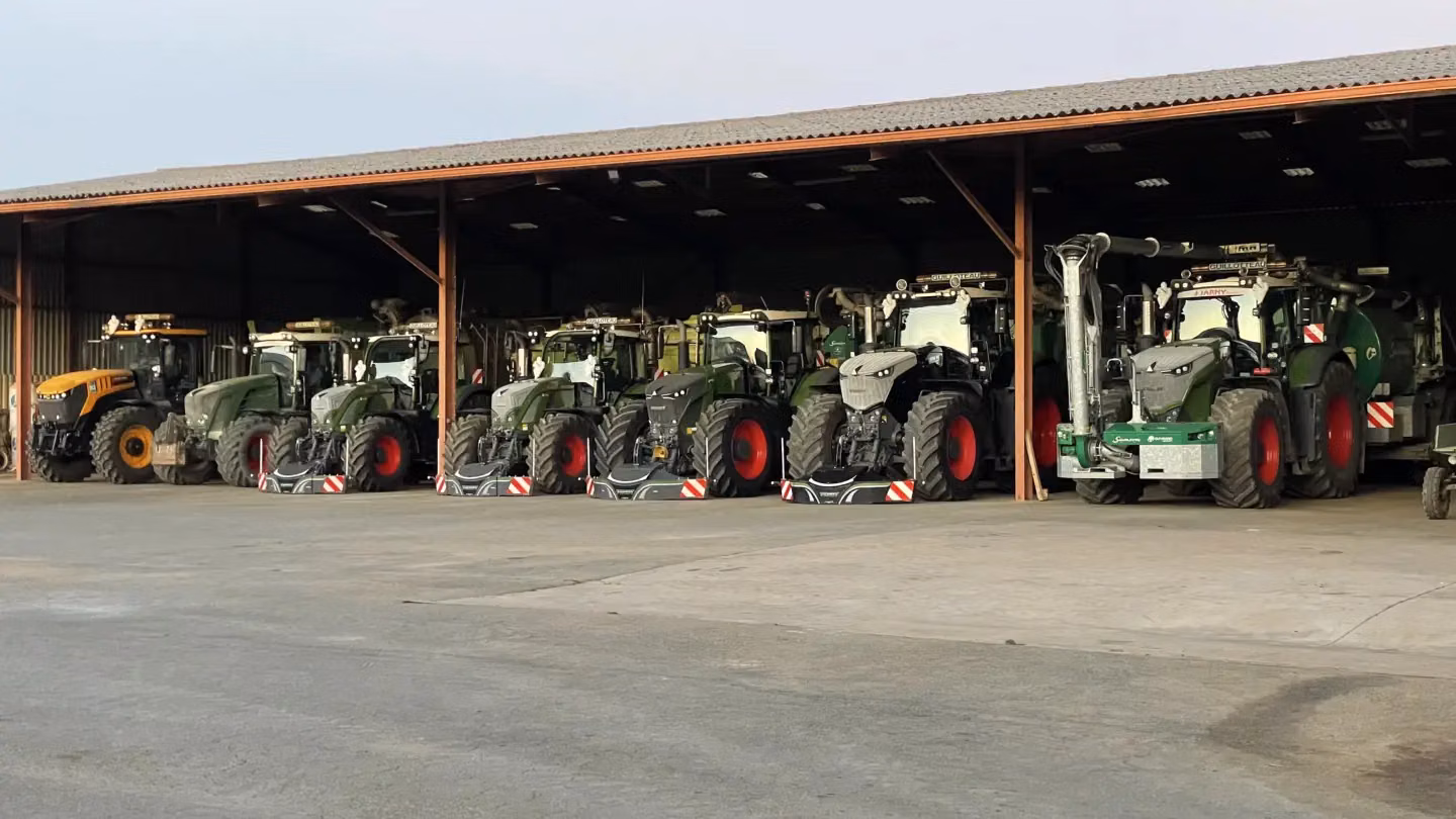 Several tractors with attached front weight bumpers are parked in a row inside an open-sided shed