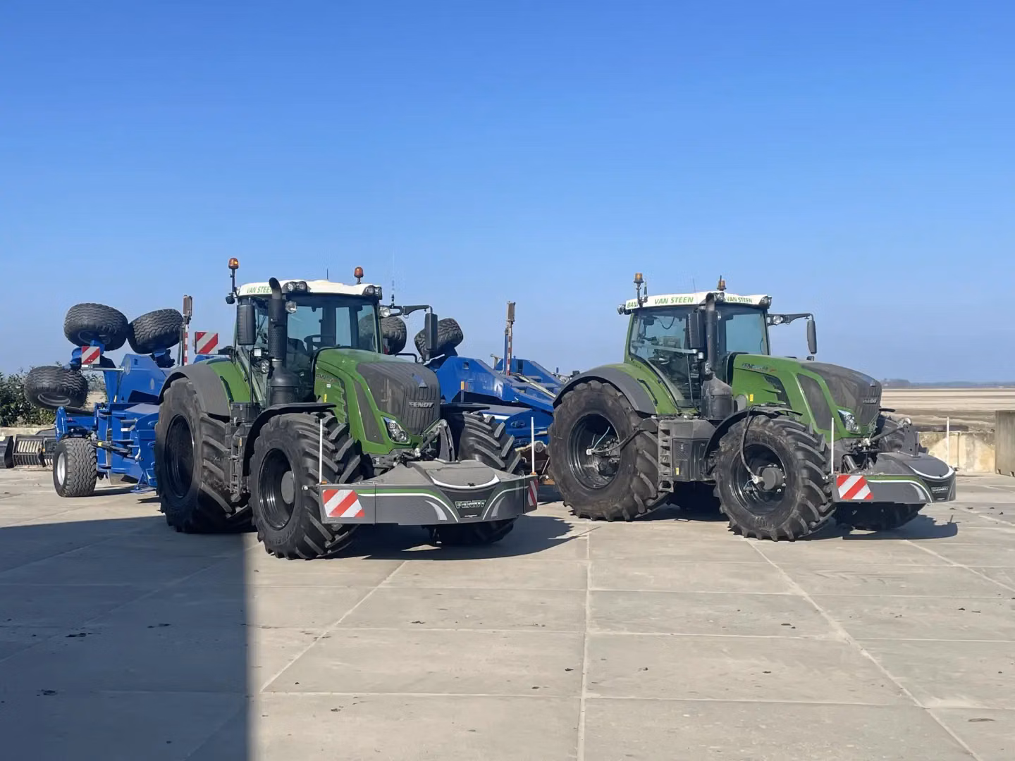 Two green tractors with large front weight bumpers are parked side by side on a concrete surface in sunlight