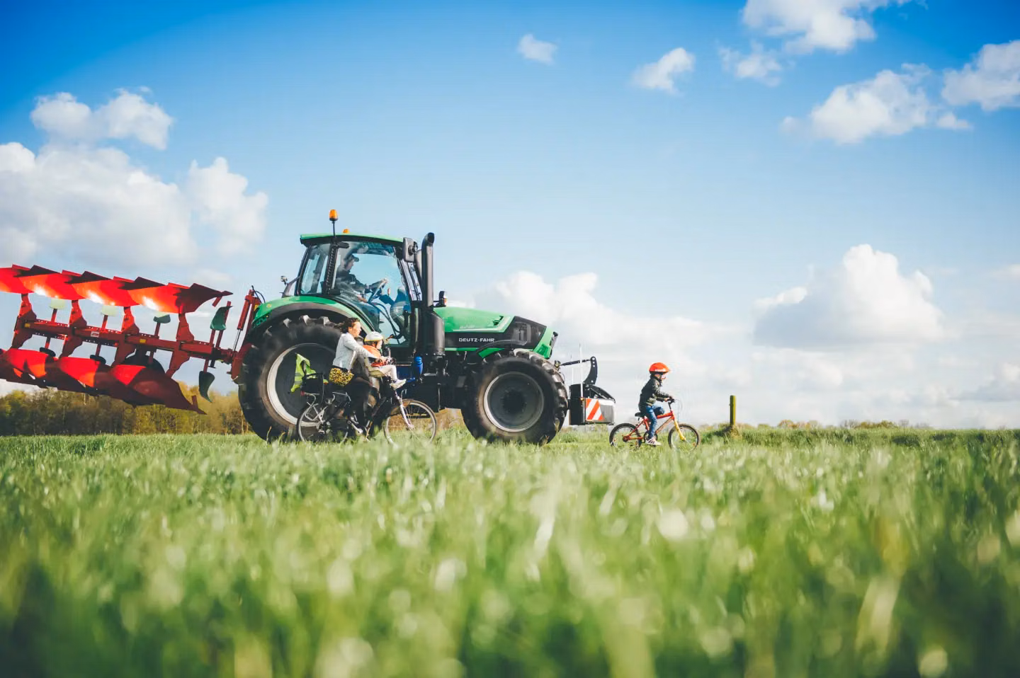 A green tractor with a front weight bumper and red tillage equipment in a grassy field, near a woman and children on bicycles