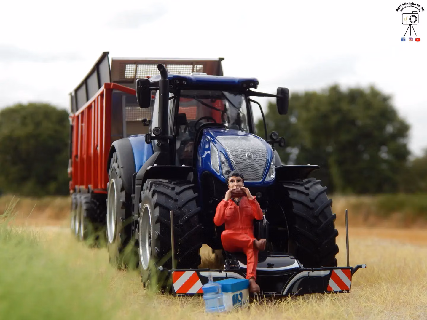 A blue tractor with a front weight bumper and attached trailer, with a figurine in red overalls sitting on the bumper