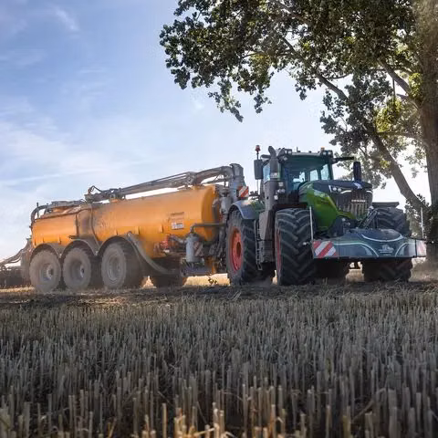 A tractor with a front weight bumper is pulling a large orange tank trailer across a harvested field.