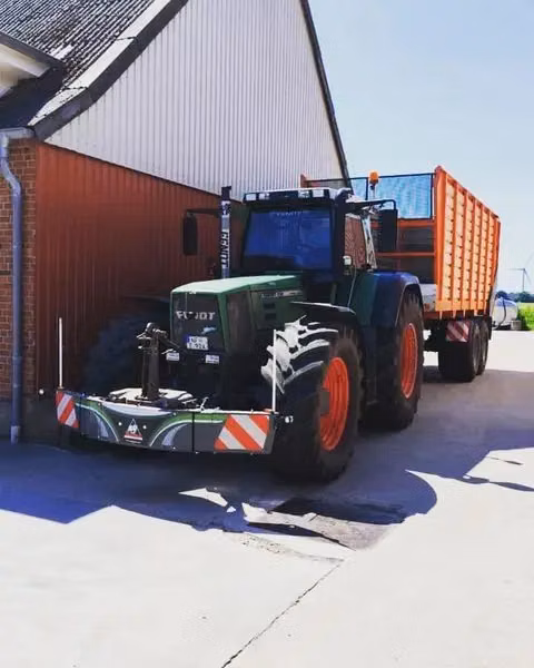 A tractor with a front weight bumper and a large orange trailer is parked beside a building on a sunny day