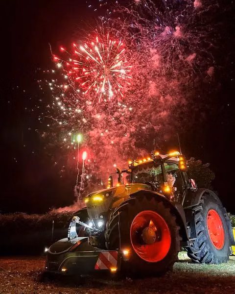 Tractor with a front weight bumper parked on grass at night with bright fireworks in the sky above
