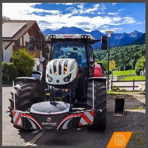 A tractor with a front weight bumper attachment is parked on a road with mountains and houses in the background