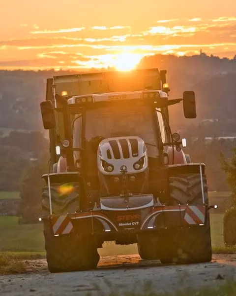 A tractor with a front weight bumper drives on a rural path at sunset with fields and hills in the background