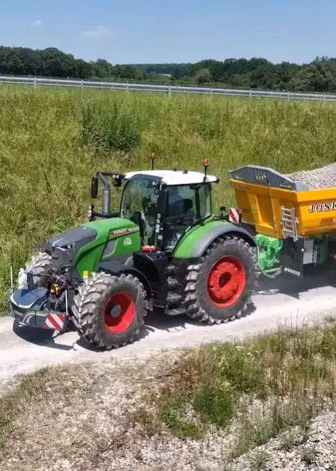 Green tractor with a front weight bumper and LED lights towing a yellow gravel trailer on a rural path